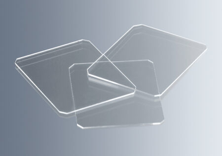 Three Haemocytometer Cover Glasses (20 x 26 x 0.4mm) overlap with beveled corners on a reflective surface against a light gradient background.