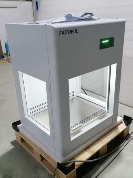 A Laminar Flow Cabinet, Vertical Class 100 with Colour screen LCD controller for Operator and Environment Protection sits on a wooden pallet, featuring glass sides and an illuminated electronic control panel.