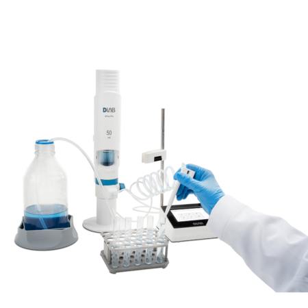 A gloved hand dispenses liquid into test tubes using laboratory equipment, including a dispenser, digital device, and a bottle with blue liquid.