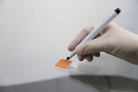 A gloved hand uses a Laboratory Marker Pen with 1mm Tip for slide labelling Black (12/Box) to write on an orange label on a white surface.