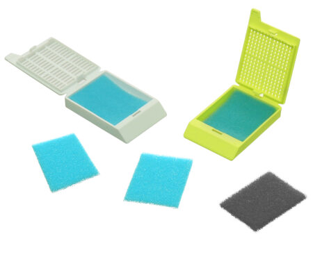 Two open cassettes, each with a blue biopsy foam pad, are shown with three loose pads—two blue and one black—in front. Product shown: biopsy foam pad (500/bag).