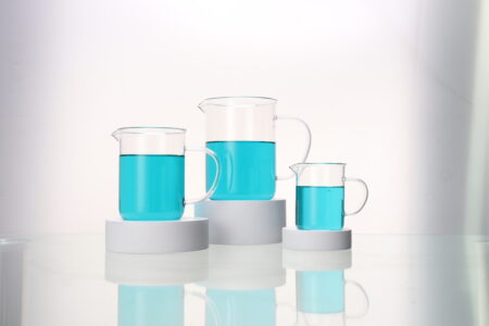 Three beaker with handle and spout in different sizes, each filled with blue liquid, are arranged on white circular platforms against a light background.