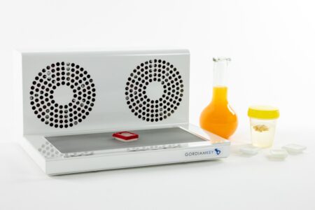 A low profile Cryoplate 4X laboratory device, designed for handling up to 27 tissue cassettes, features dual ventilation fans, a red slide, a bottle of orange liquid, a specimen jar, and four small white containers—all displayed on a white background.